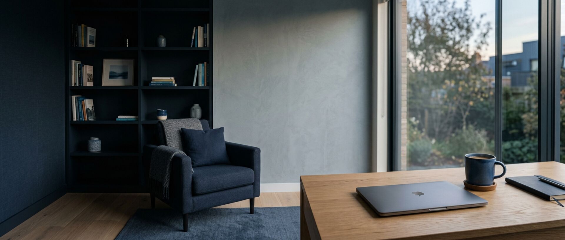 Quiet navy mental health consultation room with a shadowed reading armchair and a closed laptop on a sunlit oak table